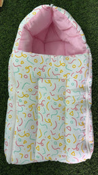 A baby footmuff or bassinet cover with a white background covered in a colorful confetti-like abstract pattern, featuring a solid pink inner lining and a zippered closure, laid flat on green grass.